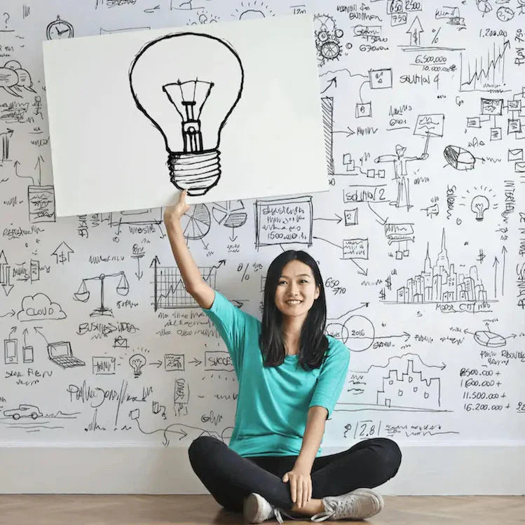 A smiling woman in a turquoise shirt sits cross-legged, holding up a sign with a drawn light bulb, against a whiteboard filled with sketches and ideas.