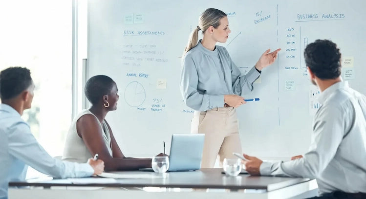 A professional lady pointing at writing on a bestboard whiteboard with a room full of people.