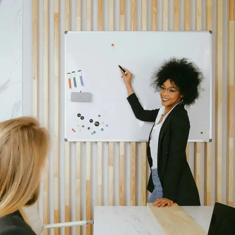 A woman in a black blazer and glasses stands in front of a whiteboard, smiling while writing with a marker. Another person with blonde hair listens attentively in a modern office setting.
