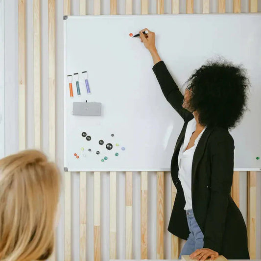 A woman in a black blazer writes on a whiteboard while another person watches, holding a tablet.