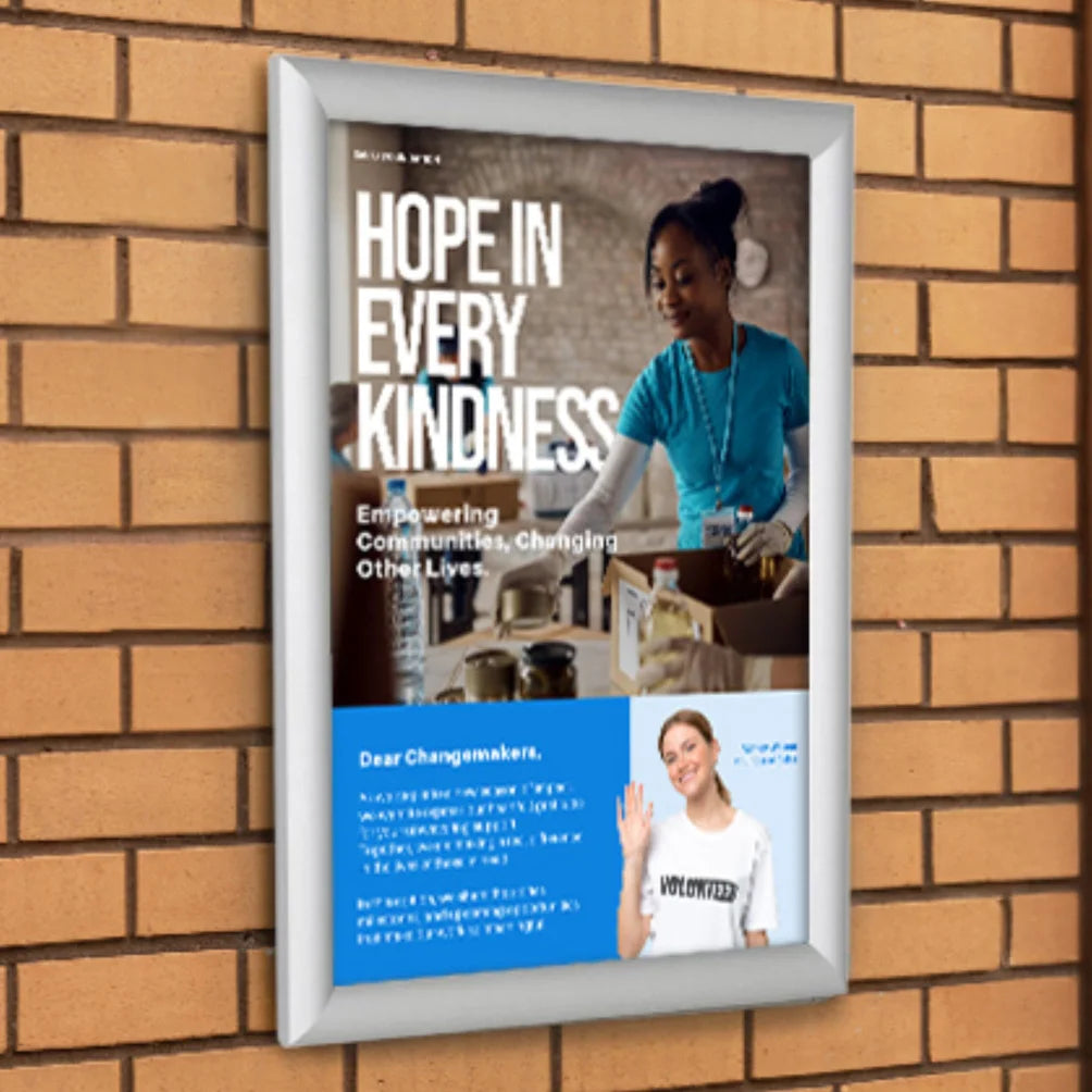 A Snap framed poster with the text 'HOPE IN EVERY KINDNESS' and a photo of a woman volunteering, mounted against a tan brick wall.