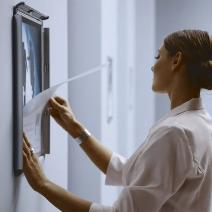 A woman in a white uniform inserts a document into a wall-mounted snap frame noticeboard with a hinged screen door.