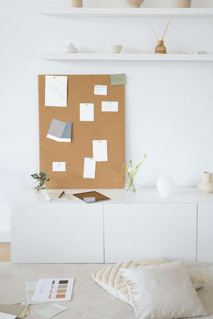 A corkboard with pinned notes and color swatches leans against a white wall, surrounded by minimalistic decor, a cabinet, and design materials on the floor.
