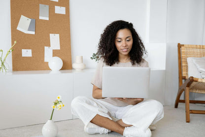 A woman with curly hair sits cross-legged on the floor, using a laptop in a bright, minimalistic room with a corkboard and decor behind her.