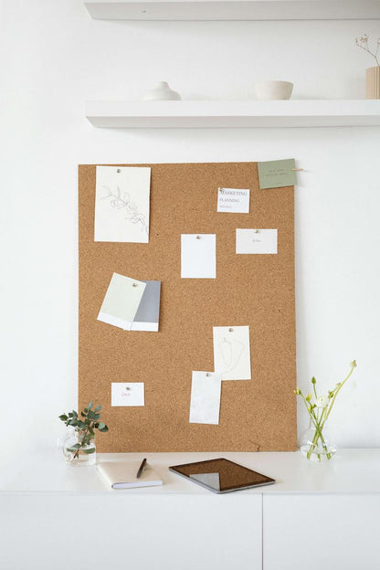 A corkboard with various pinned notes and sketches, placed on a white cabinet with a notebook, pen, and tablet in front; minimalistic decor with small vases and shelves in the background.